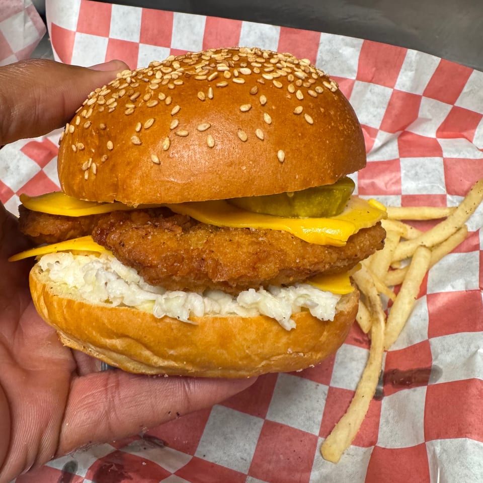 Hand holding a fried fish sandwich with cheese, pickles, and coleslaw, served with fries.