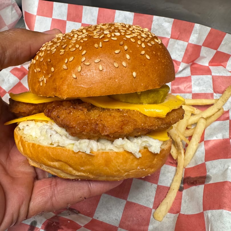 Hand holding a fried fish sandwich with cheese, pickles, and coleslaw, served with fries.