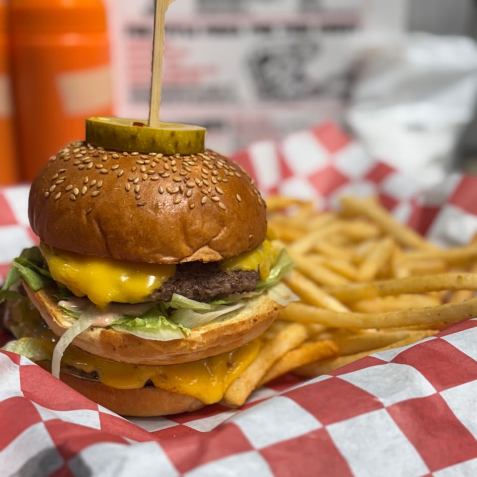 Double cheeseburger with lettuce, sauce, and a pickle on top, served with fries in a red checkered basket.