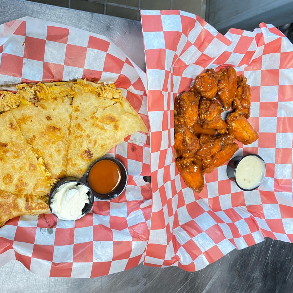A quesadilla with sour cream and hot sauce, and a basket of buffalo wings with ranch dressing.