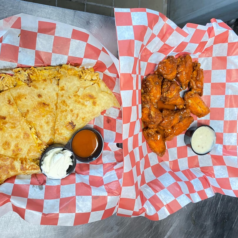 A quesadilla with sour cream and hot sauce, and a basket of buffalo wings with ranch dressing.