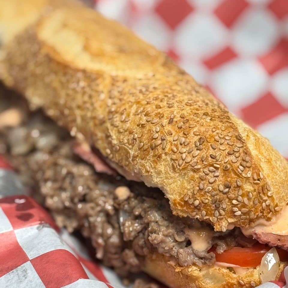 Close-up of a Pitchers Pub cheesesteak sandwich with tomatoes, onions, and sauce on a sesame seed bun.