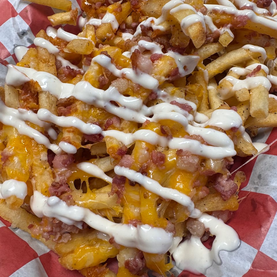 Close-up of loaded fries with melted cheese, bacon bits, and white sauce in a red and white checkered basket.