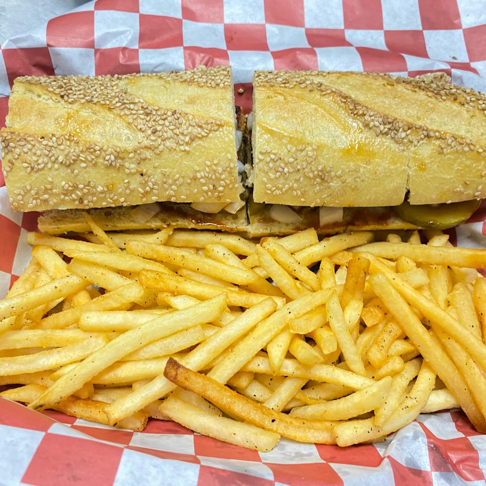 A Pitchers Pub cheesesteak sandwich, cut in half, with a side of golden french fries in a red and white checkered basket.